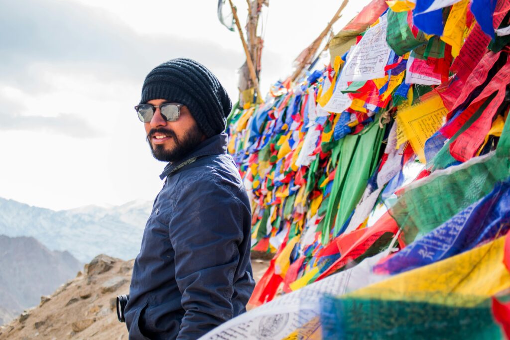 Man in sunglasses and beanie with colorful prayer flags against a mountainous backdrop.